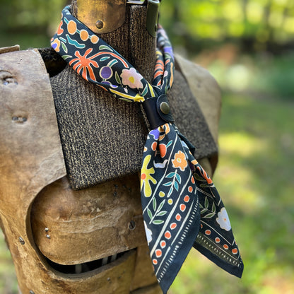 A patterned cotton bandana scarf with a black base and colorful floral motifs, styled on a rustic wooden armor display against a blurred outdoor background.