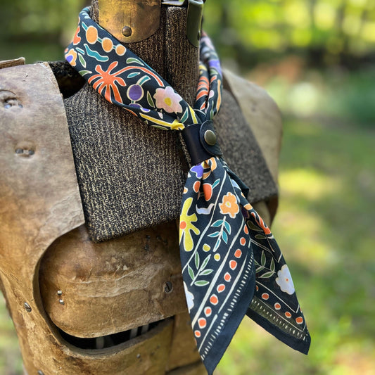 A patterned cotton bandana scarf with a black base and colorful floral motifs, styled on a rustic wooden armor display against a blurred outdoor background.
