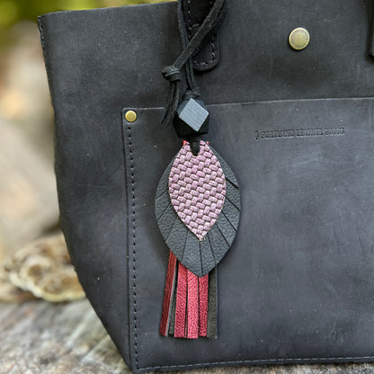 A close-up of a black leather tote bag featuring a decorative hanging charm with a pink and black leaf design and metallic tassels, set against a blurred outdoor background.
