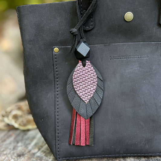 A close-up of a black leather tote bag featuring a decorative hanging charm with a pink and black leaf design and metallic tassels, set against a blurred outdoor background.