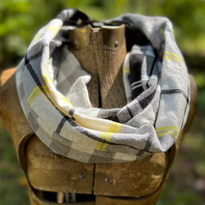 A plaid infinity scarf in white, gray, black, and yellow, draped on a rustic wooden mannequin torso with a blurred green outdoor background.
