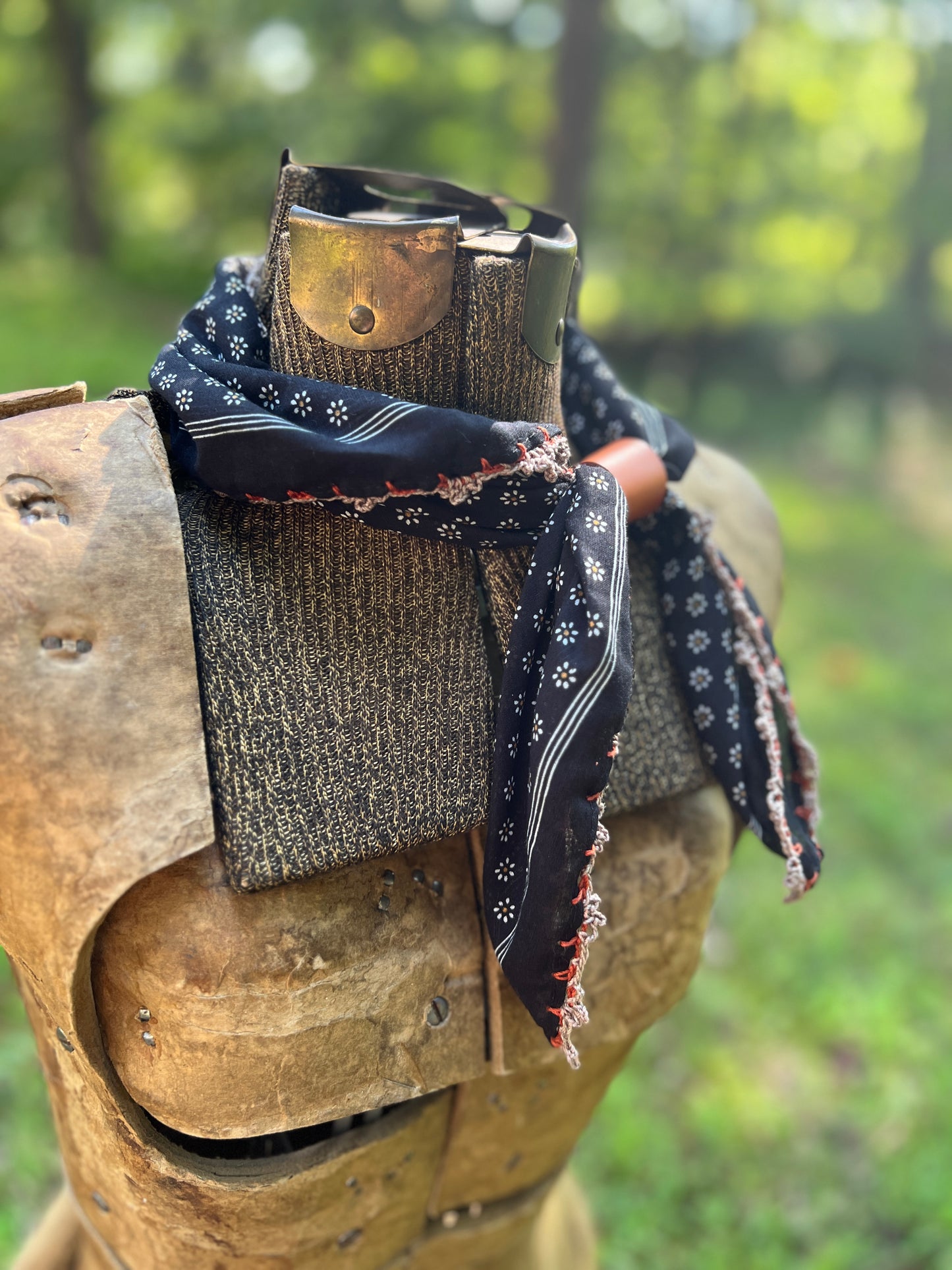 A black bandana with white and red patterns is styled around the neck of a medieval-style wooden armor plate, set against a blurred green outdoor background.