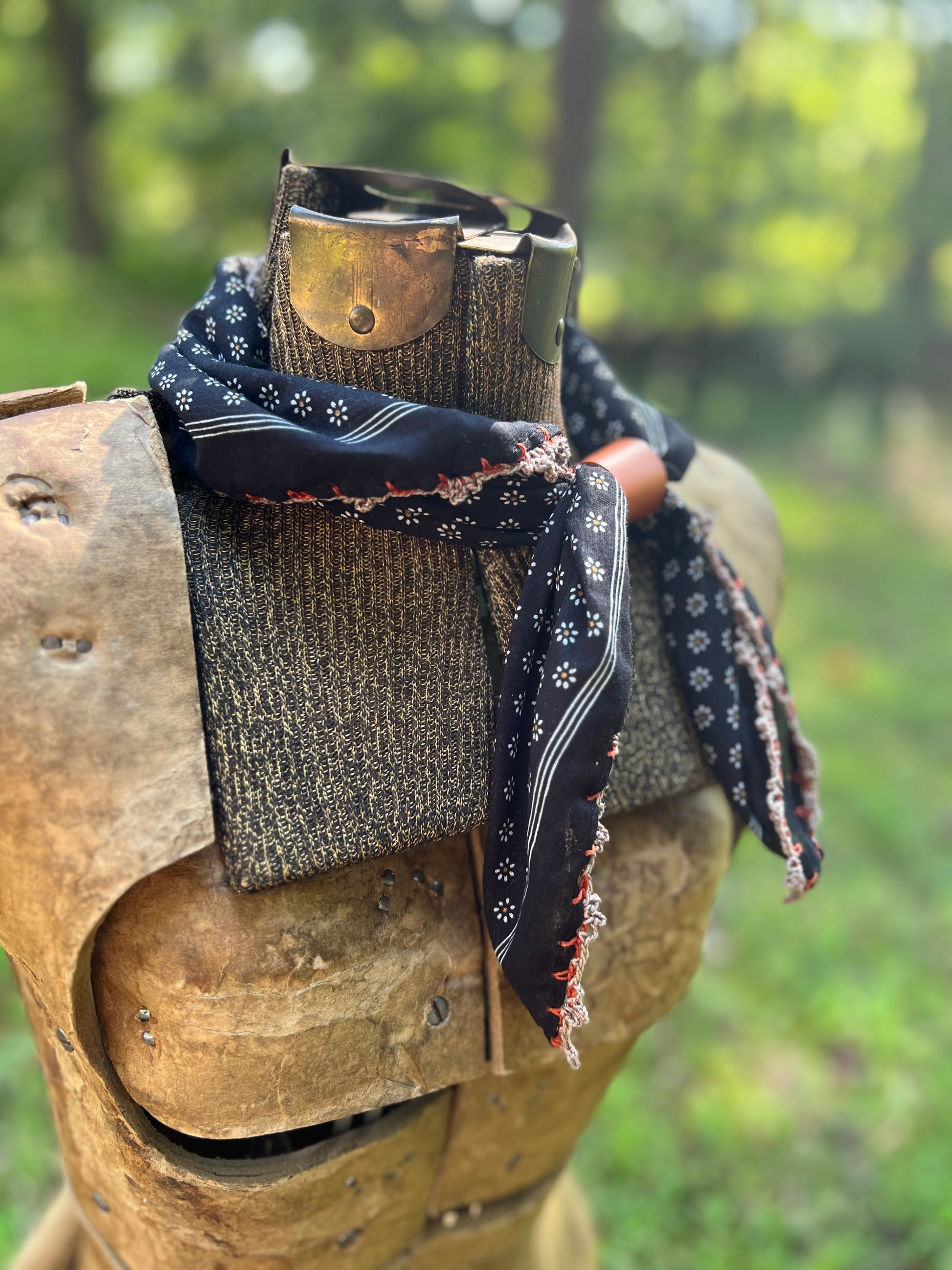 A black bandana with white and red patterns is styled around the neck of a medieval-style wooden armor plate, set against a blurred green outdoor background.