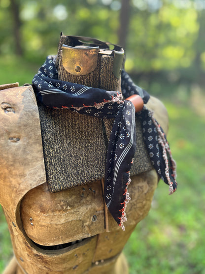 A black bandana with white and red patterns is styled around the neck of a medieval-style wooden armor plate, set against a blurred green outdoor background.