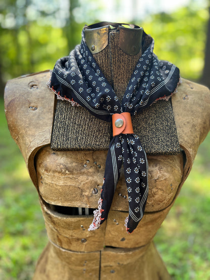 A black patterned scarf with white floral motifs and a rust-orange leather tie, styled on a medieval wooden armor chest piece against a blurred green forest background.