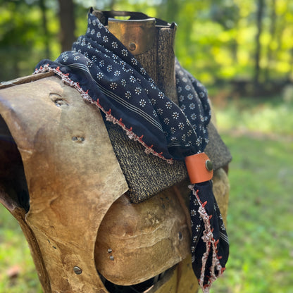A black patterned bandana with white floral motifs and red embroidered trim draped over a medieval-style wooden armor display in an outdoor garden setting.
