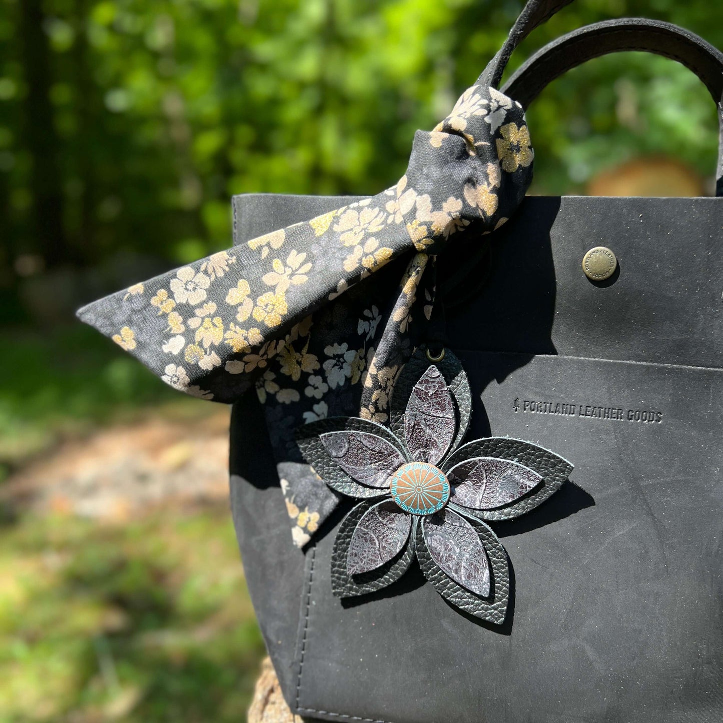 Floral-patterned bow and leather flower bag charm on a black leather bag with blurred green background