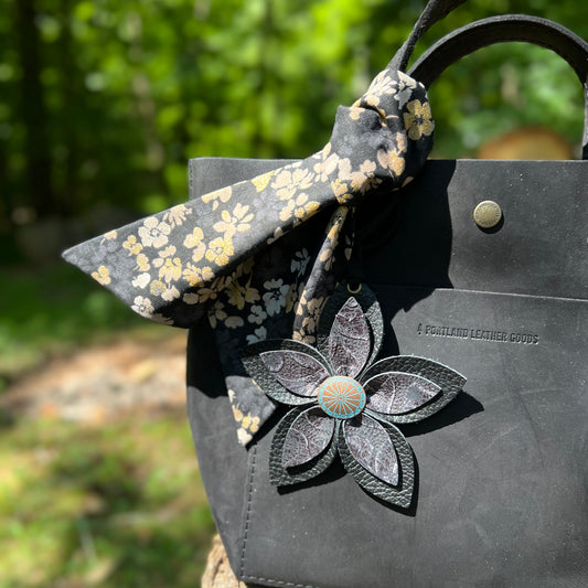 Floral-patterned bow and decorative flower on a black leather bag with blurred green background