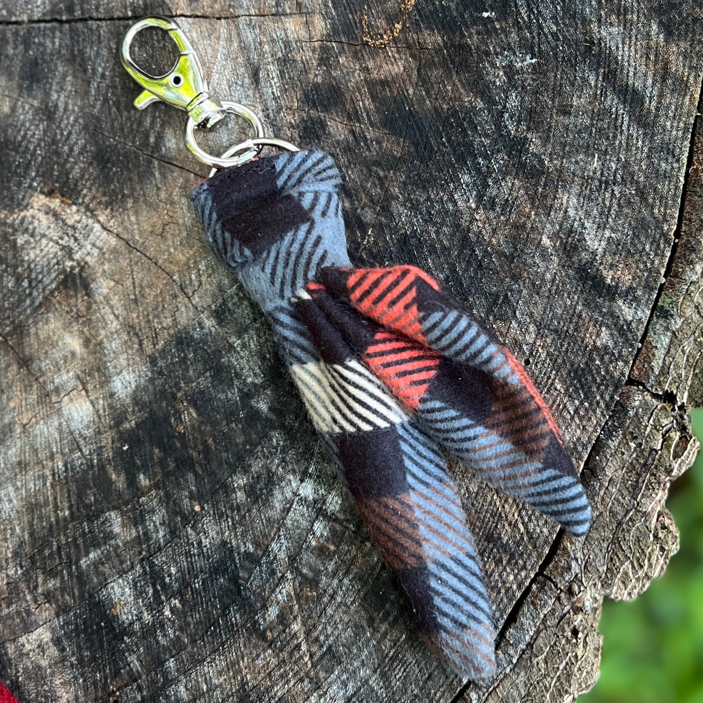 A small fabric keychain or bag charm with a plaid pattern in black, gray, red, and white, attached to a silver metal clasp, resting on a textured tree stump.