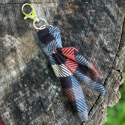 A small fabric keychain or bag charm with a plaid pattern in black, gray, red, and white, attached to a silver metal clasp, resting on a textured tree stump.