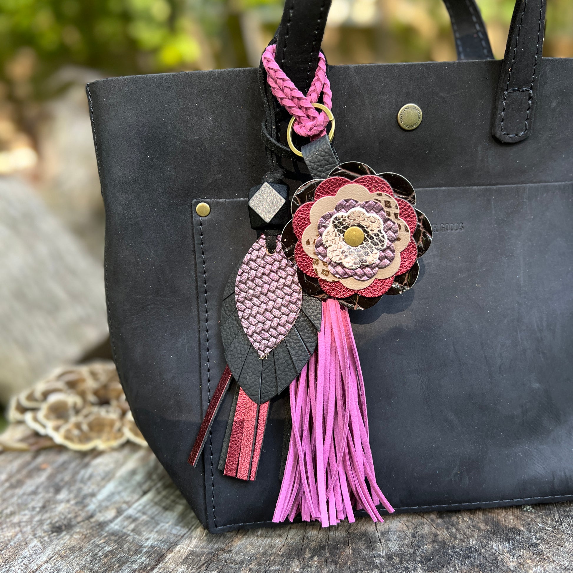 A black leather tote bag with a decorative hanging charm featuring a pink braided strap, a large layered flower in pink, burgundy, and beige tones, and a pink tassel, shown outdoors on a rustic wooden surface.
