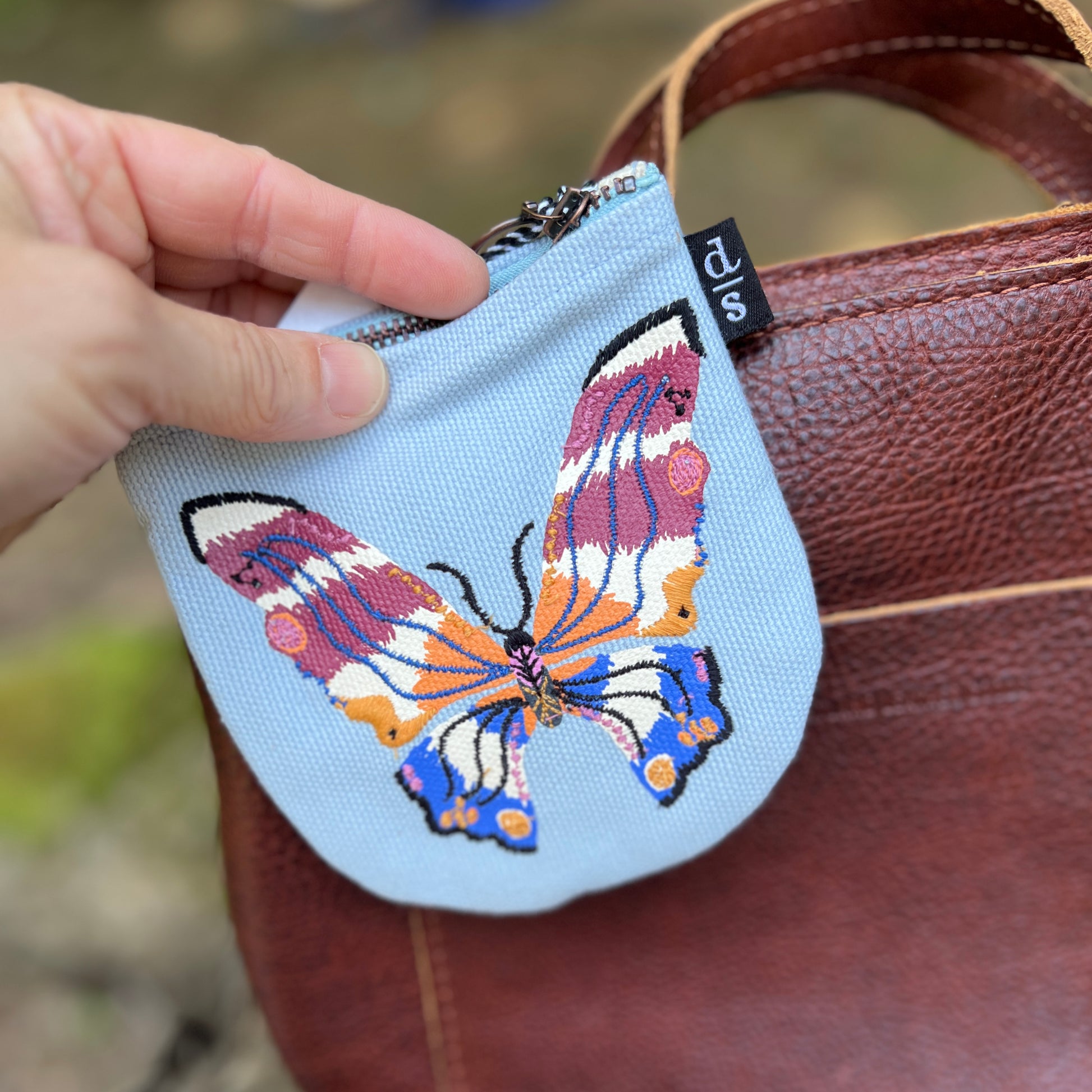 Small pouch with a butterfly design held by a hand against a brown leather bag.