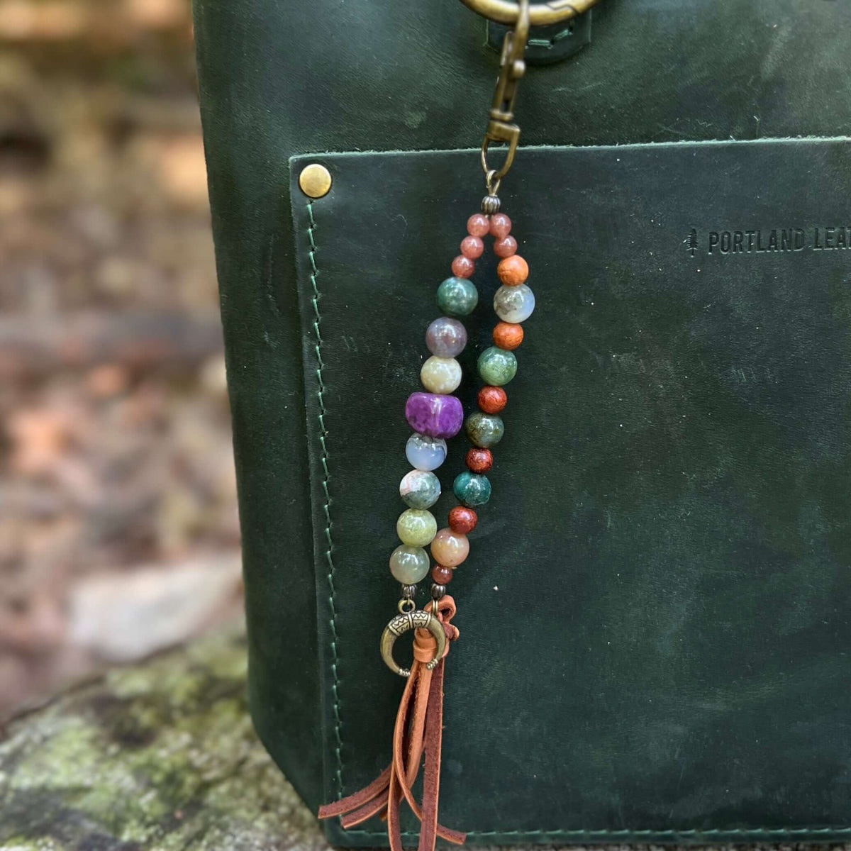 A beaded leather keychain charm with multicolored gemstone beads and a brown leather tassel, hanging from a dark green leather bag outdoors.