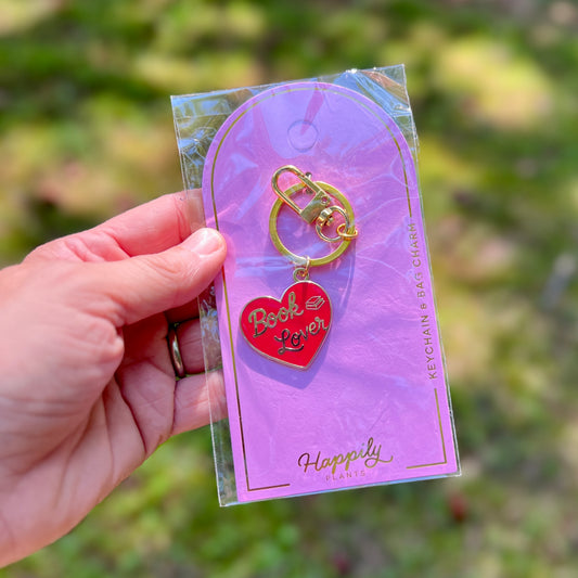 A hand holds a red heart-shaped keychain charm with the words 'Book Lover' in gold lettering, attached to a gold split ring and displayed on a pink backing card. The background is a blurred outdoor green landscape.