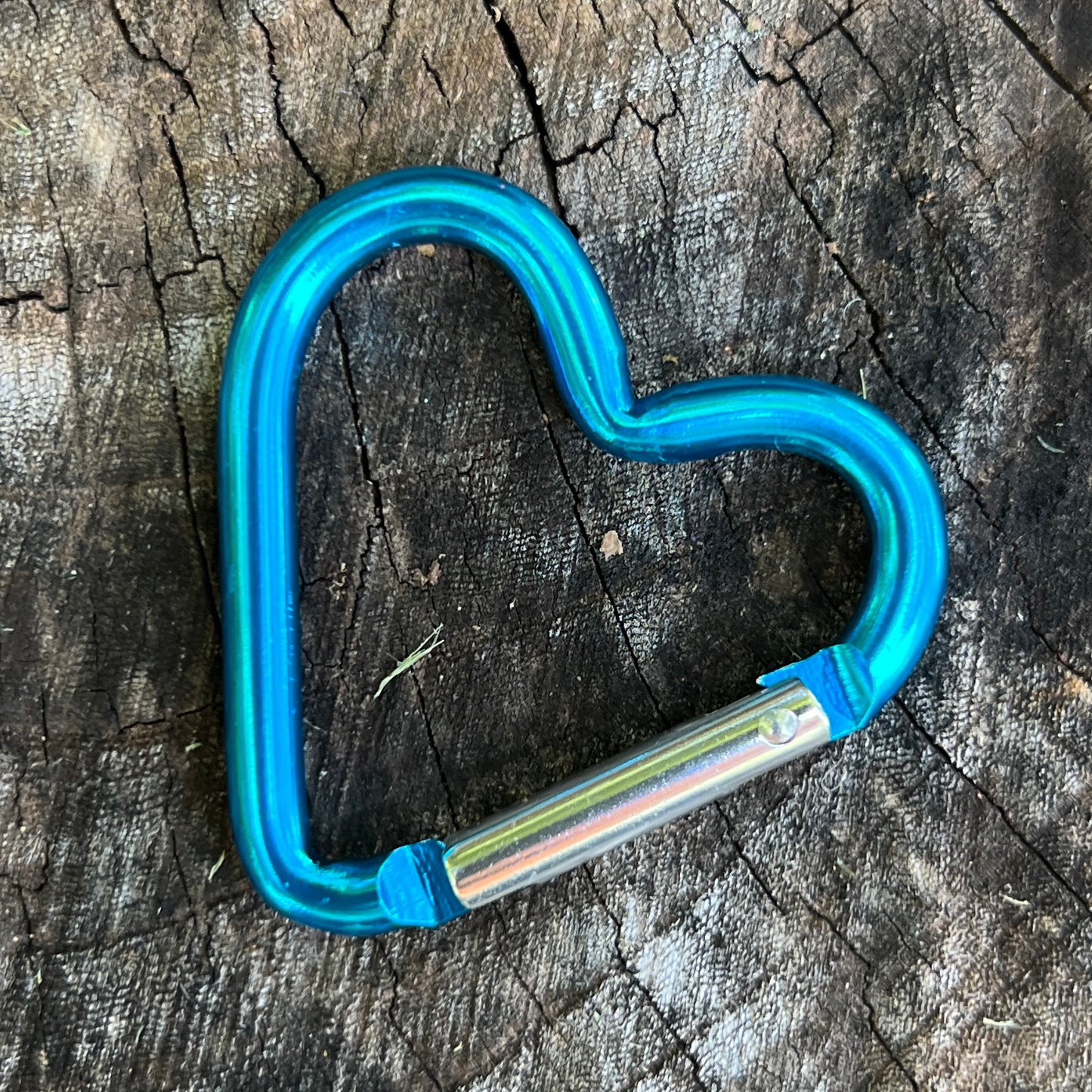 A heart-shaped carabiner with a vibrant metallic blue finish and a silver metal latch, resting on a textured, dark wood surface.