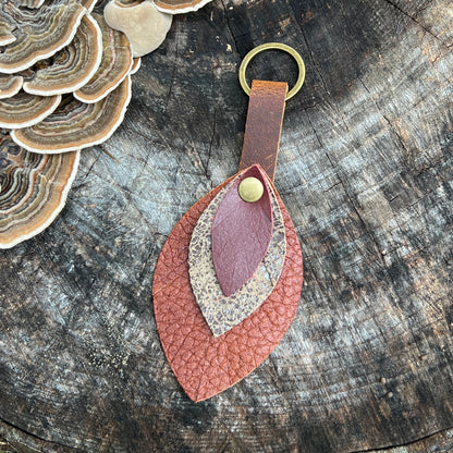 A handmade leather keychain featuring layered leaf-shaped pieces in terracotta, dusty rose, and beige, with a brass ring and snap button, displayed on a rustic wood slice background.