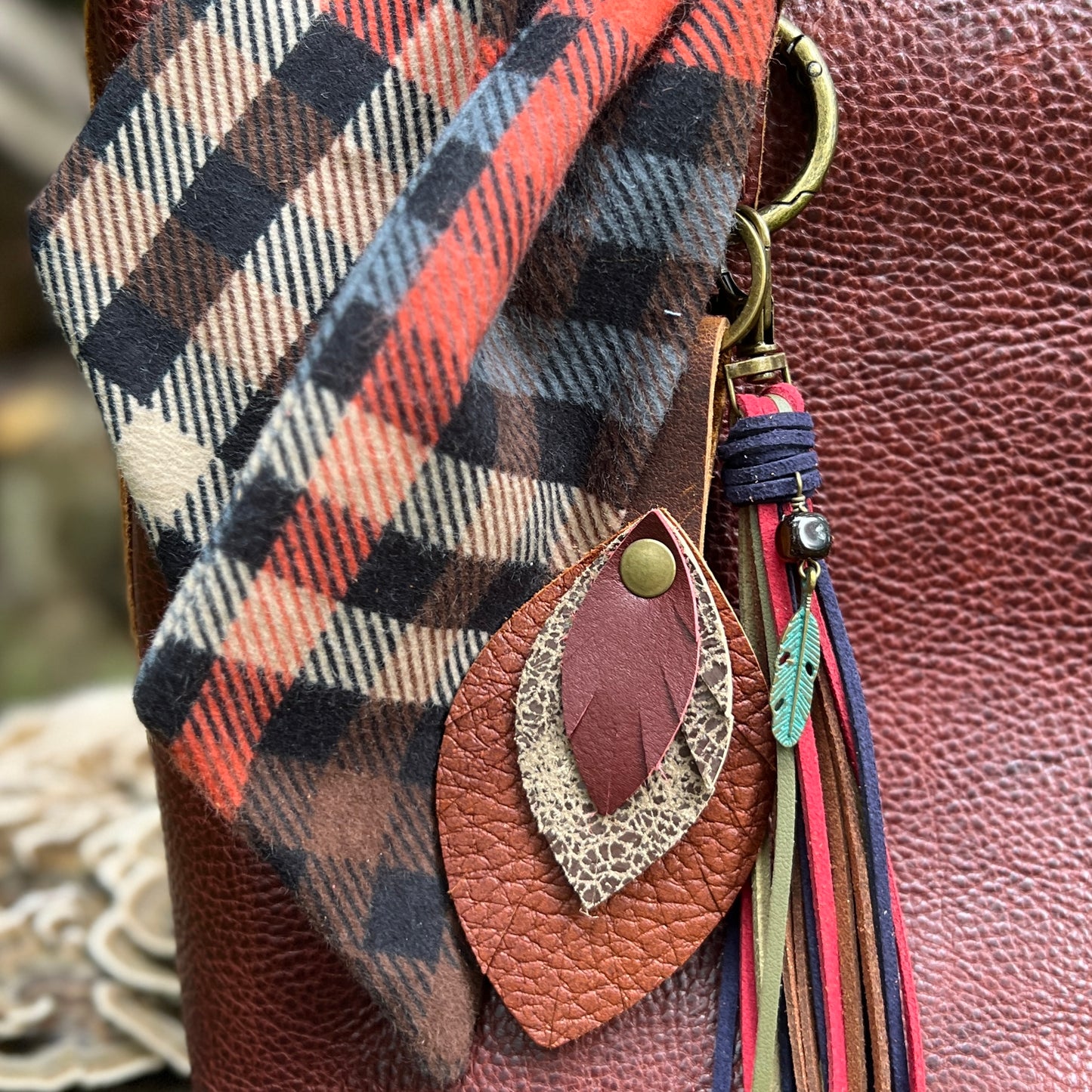 Close-up of a brown leather shoulder bag with a plaid scarf, a leaf-shaped leather charm, and a tassel keychain.