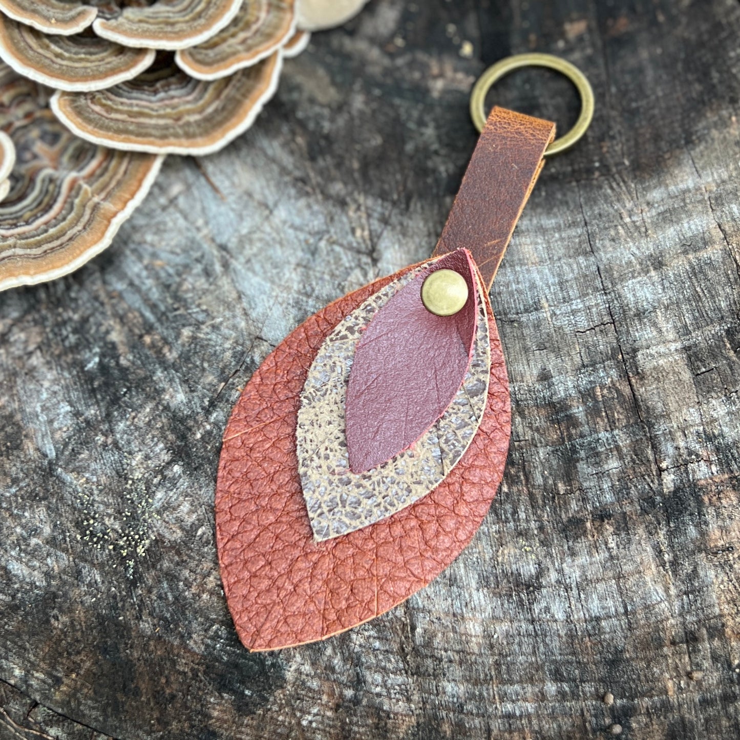 A handmade leaf-shaped leather keychain with layered textures in terracotta, dusty rose, and beige, attached to a brass key ring and resting on a rustic wood surface.