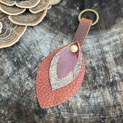 A handmade leaf-shaped leather keychain with layered textures in terracotta, dusty rose, and beige, attached to a brass key ring and resting on a rustic wood surface.