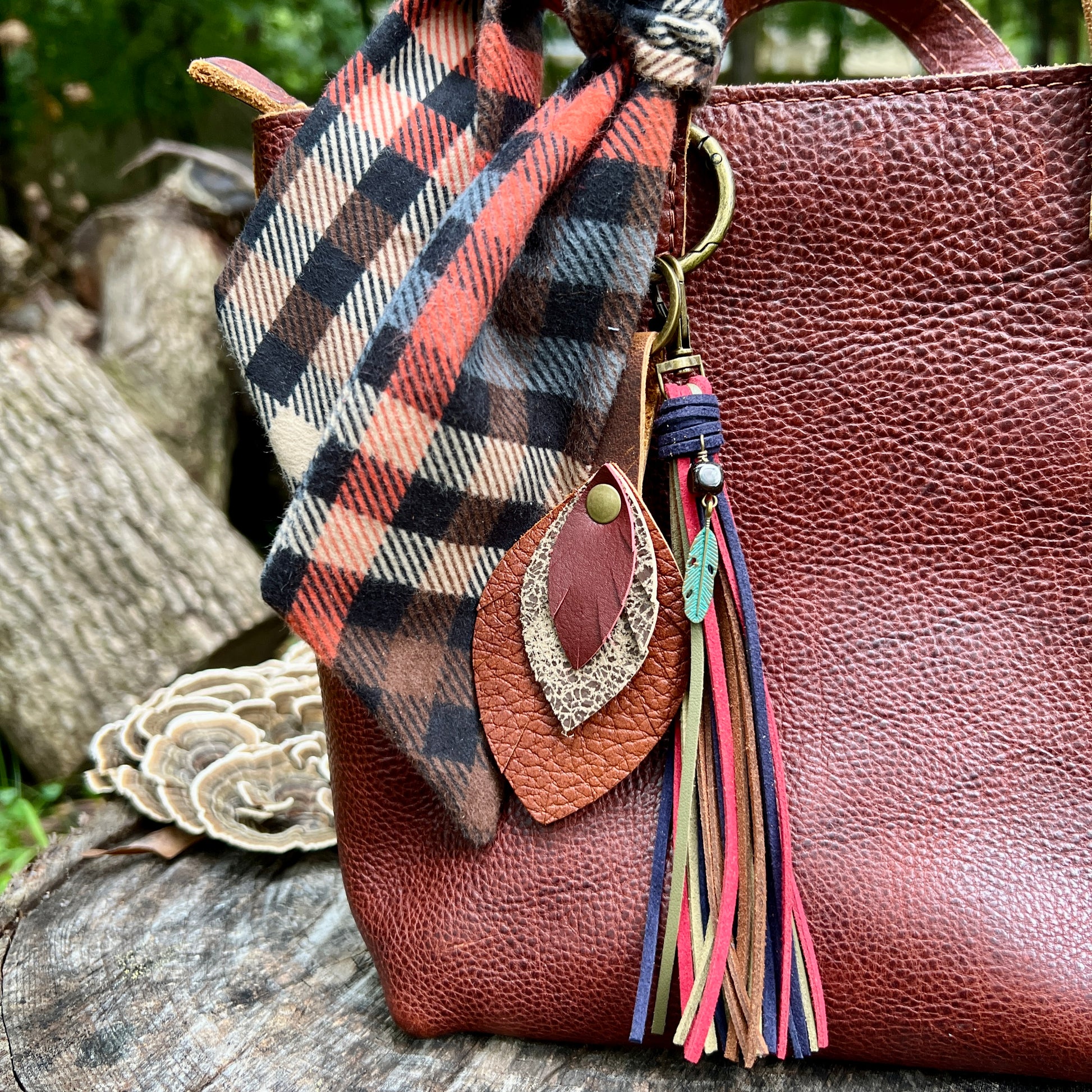 A close-up of a brown pebbled leather tote bag outdoors, styled with a rust-orange, black, and beige plaid scarf tied around the handle, and a decorative tassel keychain with a feather and colorful tassels.