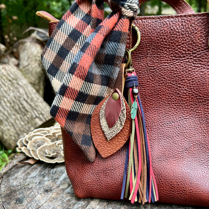 A close-up of a brown pebbled leather tote bag outdoors, styled with a rust-orange, black, and beige plaid scarf tied around the handle, and a decorative tassel keychain with a feather and colorful tassels.