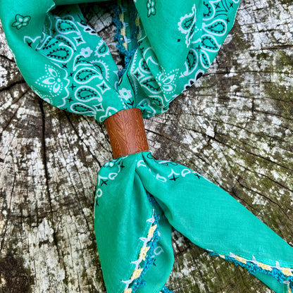 A green paisley bandana tie-on scarf with a brown leather wrap, displayed on a weathered wood surface.