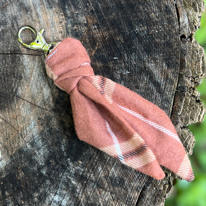 A small fabric keychain charm made from brown plaid material with a gold-tone lobster clasp, photographed resting on a textured tree stump outdoors.