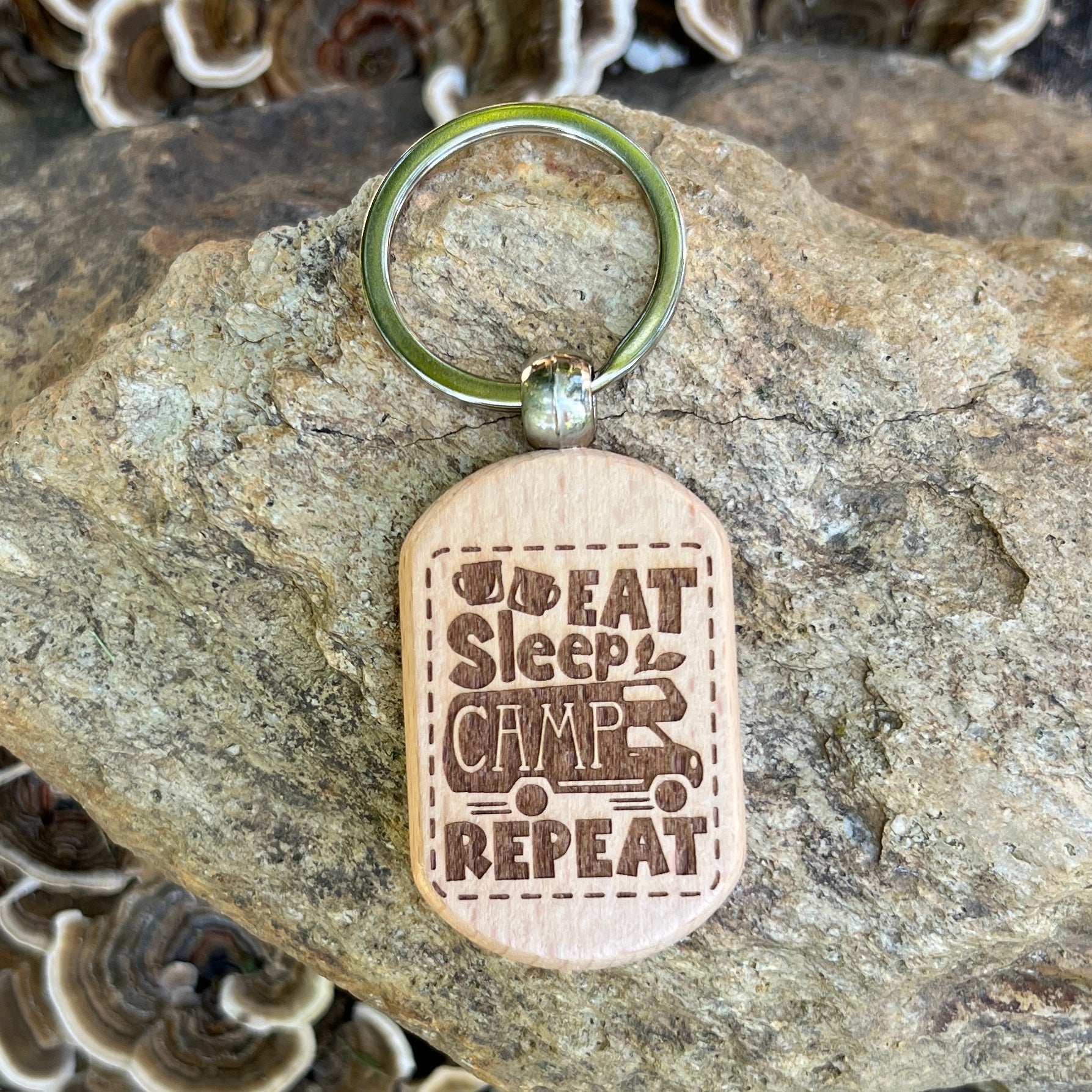 A wooden dog-tag style keychain with a metal key ring, photographed resting on a large rock. The engraved wood reads “I Eat Sleep Camp Repeat” with a small camping truck graphic.
