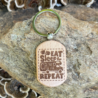 A wooden dog-tag style keychain with a metal key ring, photographed resting on a large rock. The engraved wood reads “I Eat Sleep Camp Repeat” with a small camping truck graphic.
