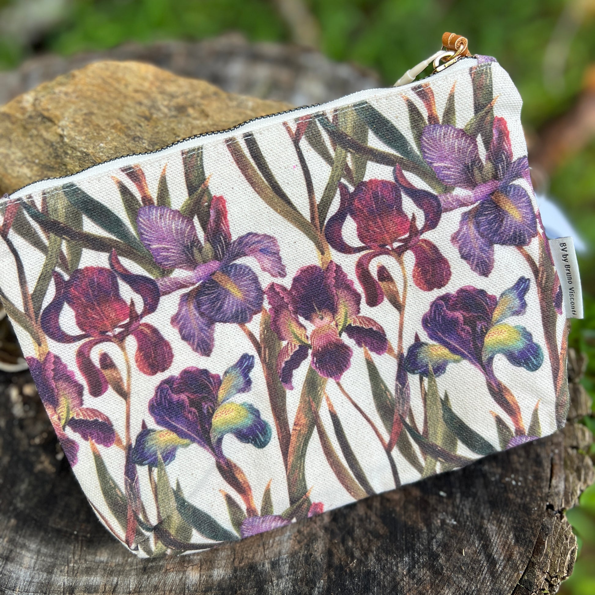 A canvas zip pouch featuring a watercolor-style print of purple irises on a white background, resting on a rustic wooden log with a blurred green outdoor background.