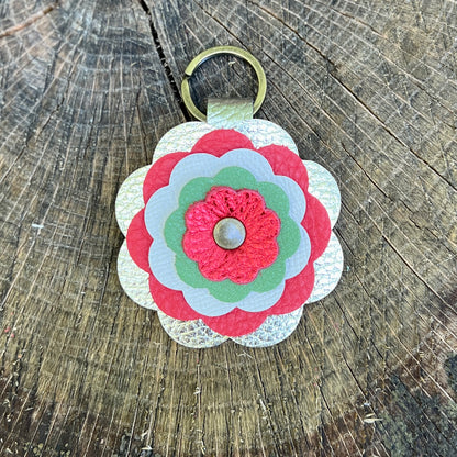 A handmade flower-shaped keychain made of layered scalloped leather in red, light green, and white, attached to a gold metal key ring, displayed on a rustic wooden surface.