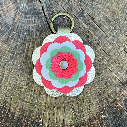 A handmade flower-shaped keychain made of layered scalloped leather in red, light green, and white, attached to a gold metal key ring, displayed on a rustic wooden surface.