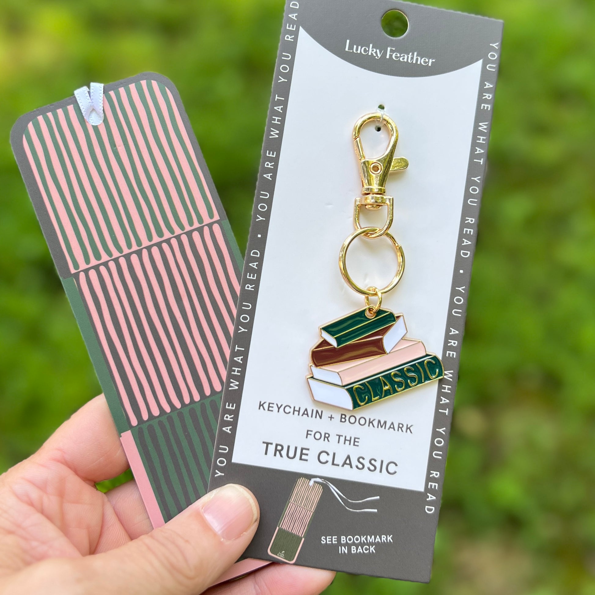 A hand holds a keychain and matching bookmark set on a branded card against a blurred green outdoor background. The keychain features stacked books with gold-tone hardware and a green enamel 'CLASSIC' section.
