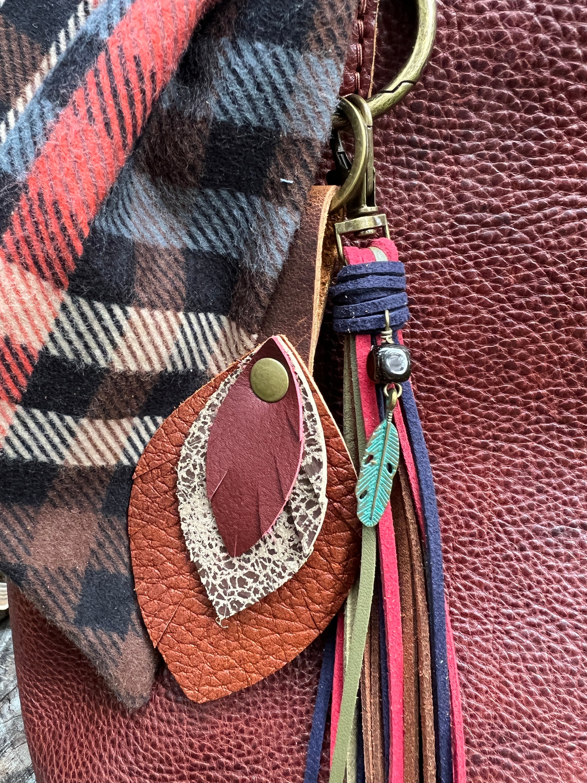 Close-up of a leather accessory with a leaf-shaped charm in brown and cream, attached to a tassel strap with pink, blue, and green cords, next to a rust-colored textured leather surface and a plaid fabric.