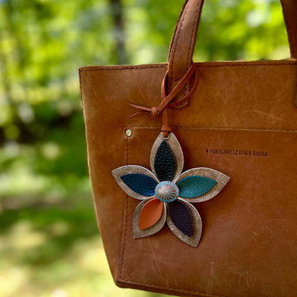 Brown leather bag with a decorative leather flower bag charm featuring blue, green, and orange colors, set against a blurred natural background.