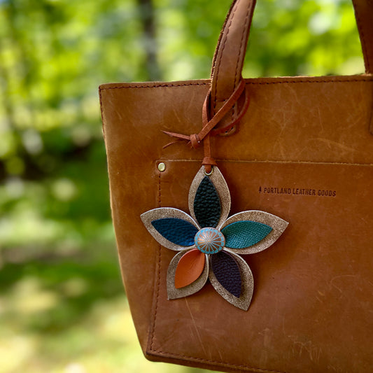 Brown leather bag with a decorative flower-shaped patch featuring blue, green, and orange colors, set against a blurred natural background.
