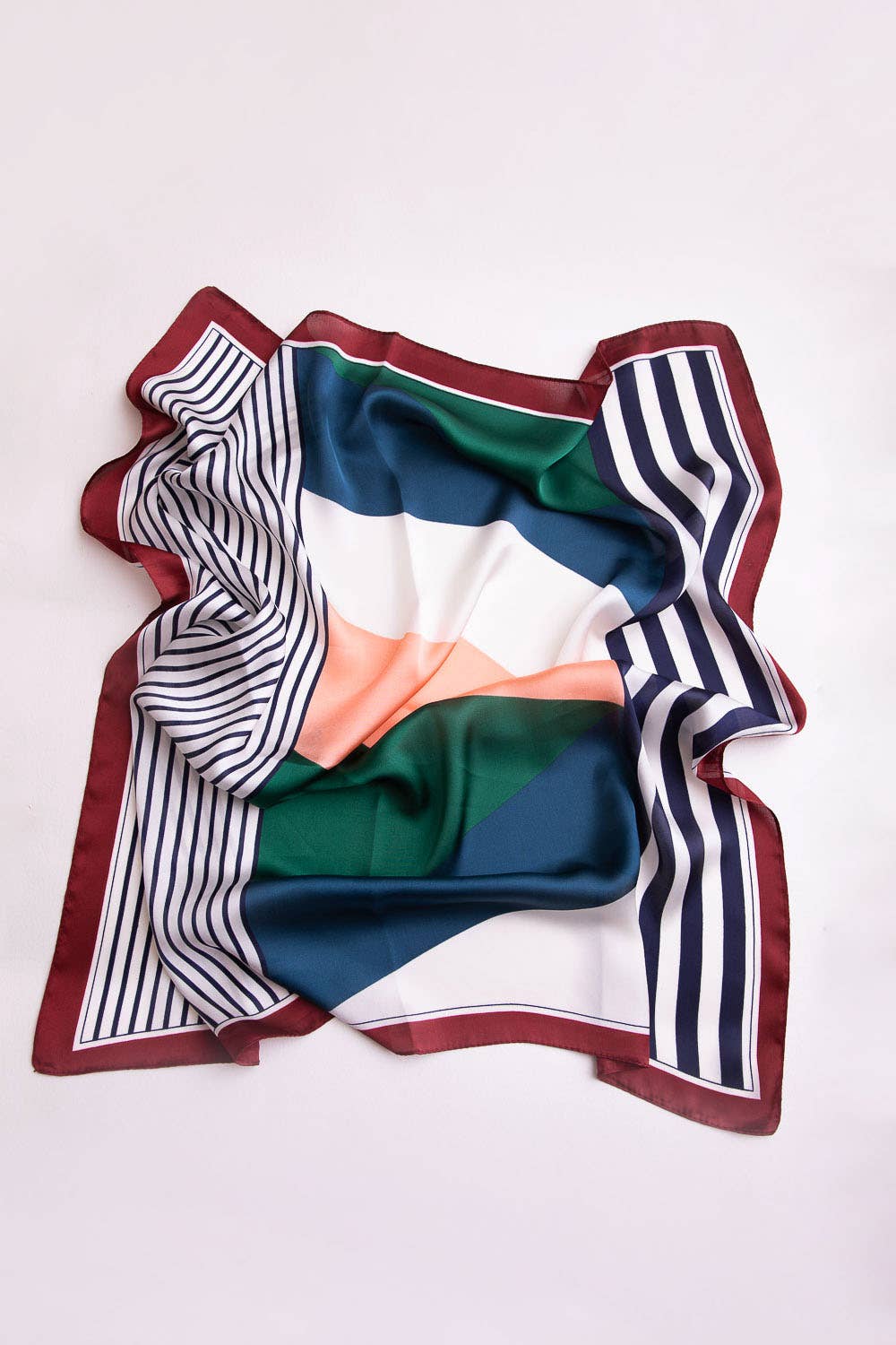 A square silk scarf with a multicolor geometric pattern, featuring stripes, solid blocks of green, blue, white, and peach, and a burgundy border, laid flat on a light off-white background.