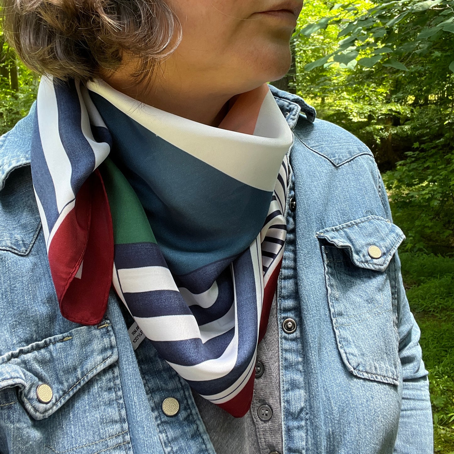 A close-up of a person outdoors wearing a striped silk scarf in blue, white, red, and green, styled under a light blue denim jacket.
