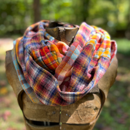 A multicolor plaid infinity scarf with orange, yellow, blue, pink, and red tones, draped on a rustic wooden mannequin torso against a blurred outdoor forest background.