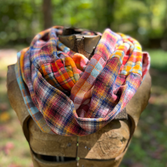 A multicolor plaid infinity scarf with orange, yellow, blue, pink, and red tones, draped on a rustic wooden mannequin torso against a blurred outdoor forest background.
