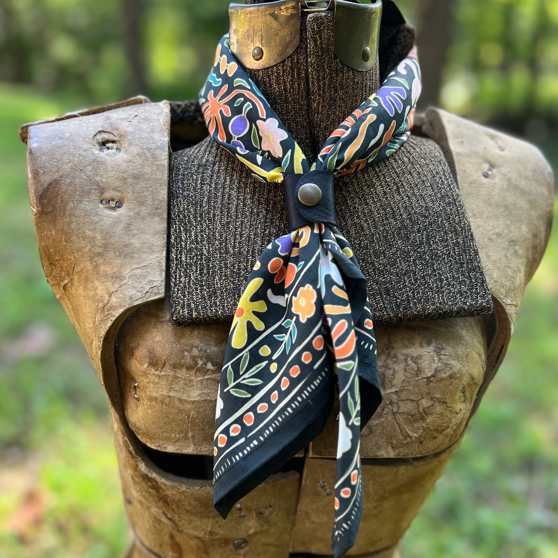 A patterned cotton bandana scarf with a black base and colorful floral motifs, styled on a medieval-style wooden armor display outdoors.