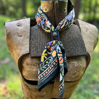 A patterned cotton bandana scarf with a black base and colorful floral motifs, styled on a medieval-style wooden armor display outdoors.