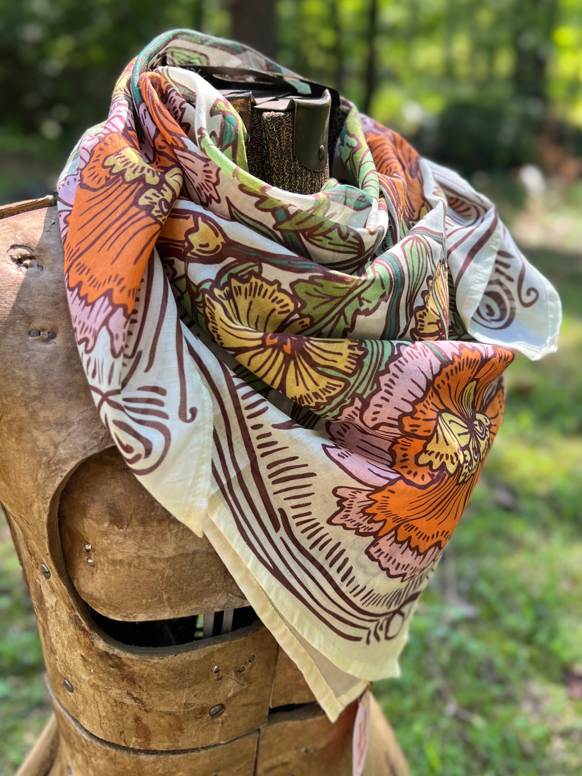 A multicolor floral scarf with orange, green, pink, and brown patterns on a cream base, draped over a rustic wooden armor display outdoors.
