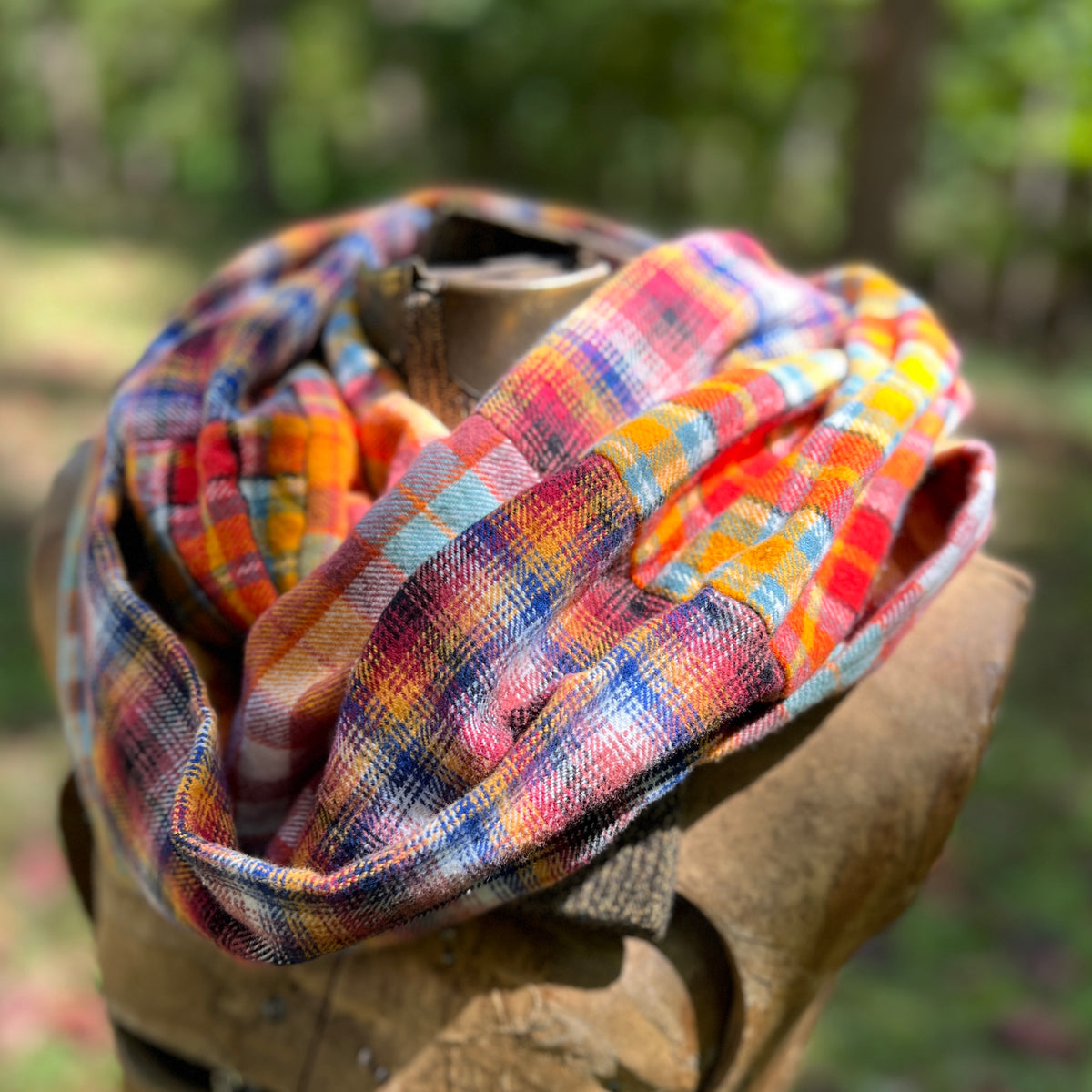 A multicolor plaid infinity scarf in red, orange, yellow, blue, and white, draped on a rustic wooden display stand outdoors with a blurred forest background.