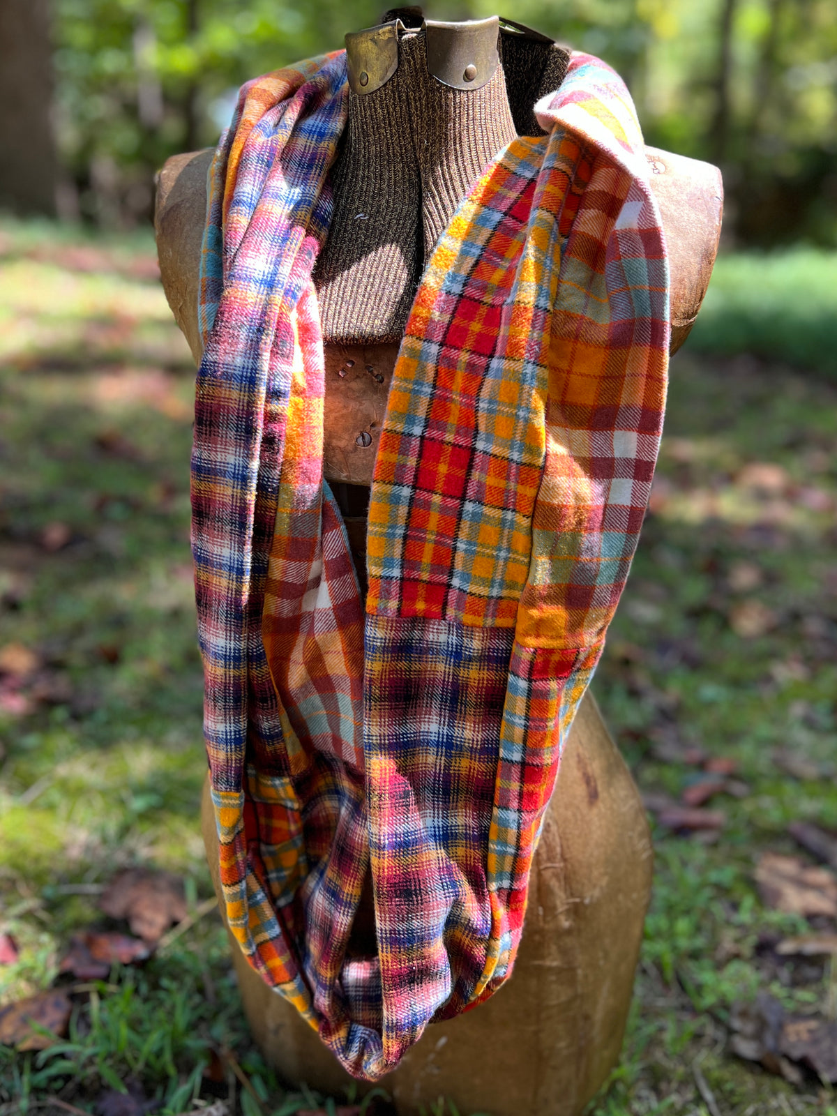 A multicolor plaid infinity scarf draped on a wooden mannequin torso outdoors in a grassy, wooded setting.
