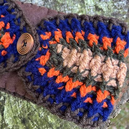 A close-up of a handmade crocheted winter headband made from chunky yarn in navy blue, bright orange, dark brown, and beige, featuring a spiral wooden button closure, set against a blurred outdoor background.