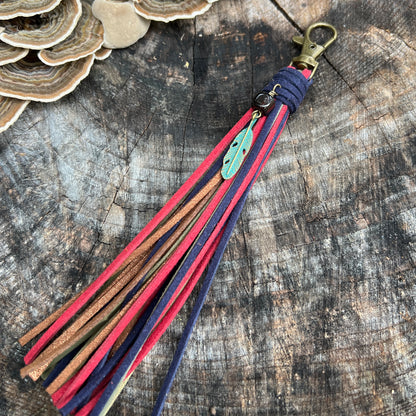 A handmade leather tassel keychain or bag charm with a brass clasp, wrapped in navy blue cord, and featuring multiple leather strands in red, brown, and navy, with a small turquoise feather charm, displayed on a rustic wood slice background.