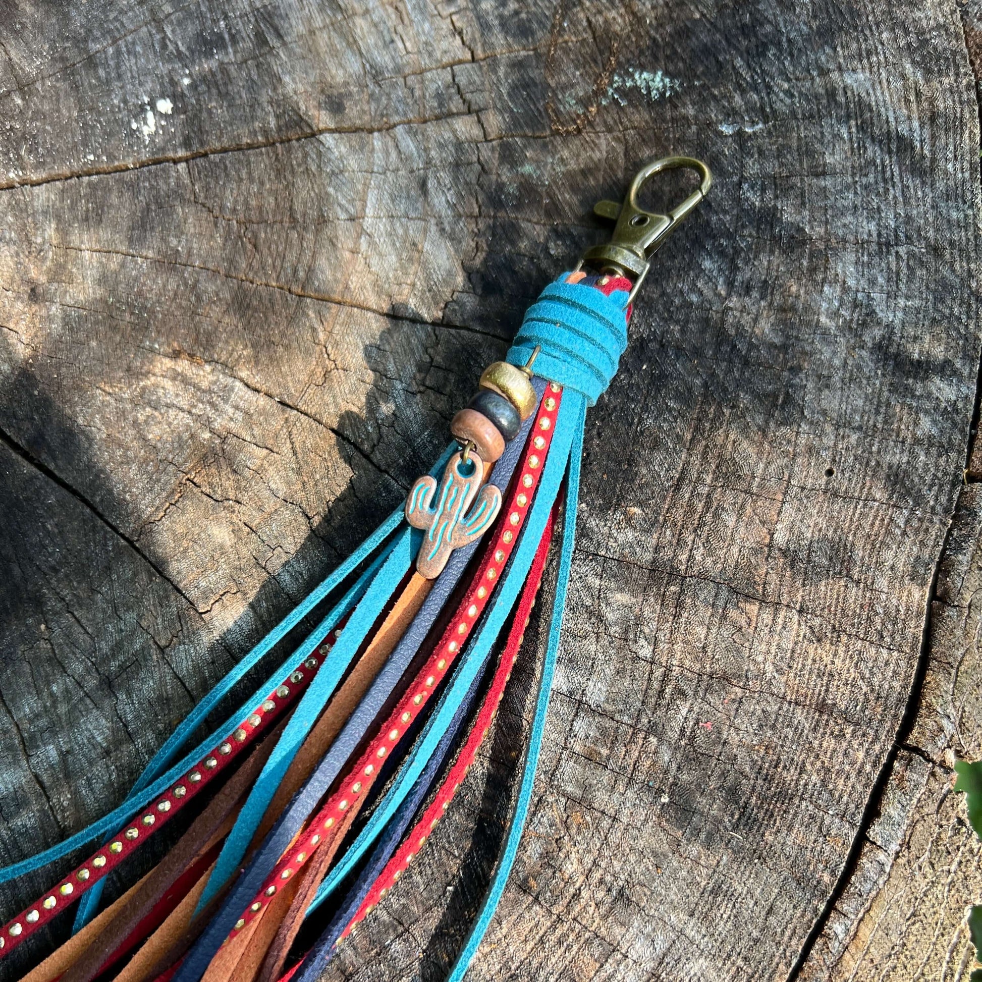 A leather tassel keychain with multiple colorful straps in turquoise, red, brown, and purple, featuring a silver cactus charm and a brass clasp, resting on a rustic wood log background.
