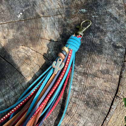 A leather tassel keychain with multiple colorful straps in turquoise, red, brown, and purple, featuring a silver cactus charm and a brass clasp, resting on a rustic wood log background.