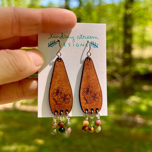 Brown teardrop earrings with floral designs and bead accents on a card held by a hand with a blurred green outdoor background.